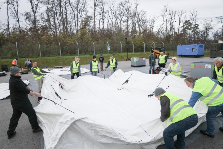 Surrogate inspectors from the in-field operations support team work in unison to position an unpacked tent and prepare it for inflation during the commissioning of the Base of Operations (BoO).