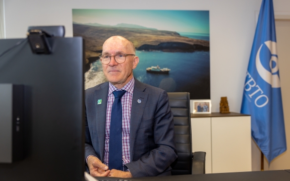 Dr Robert Floyd sits before a computer at a desk with CTBTO flag in background
